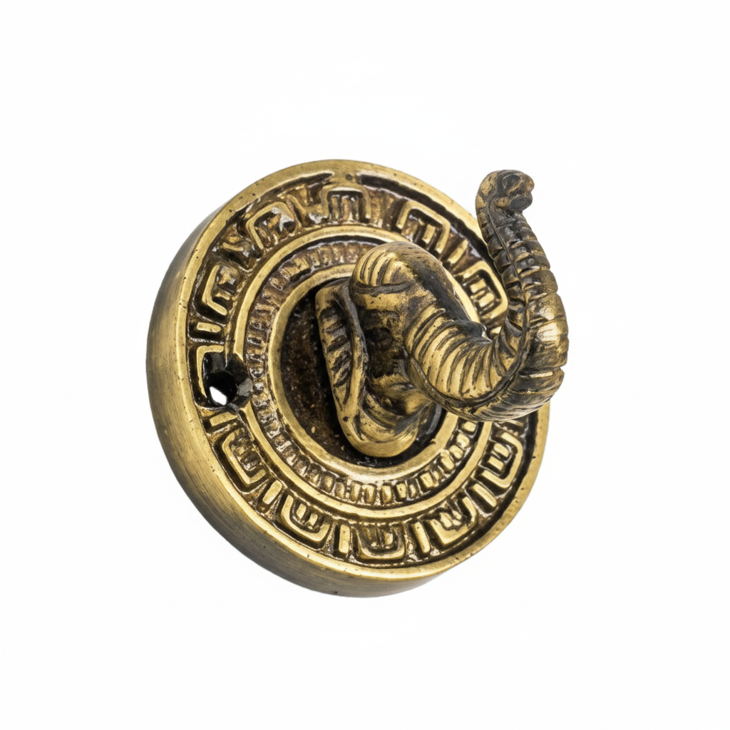 An ornate decorative wall hook featuring a detailed elephant head with an upturned trunk serving as the hook, set on a circular backplate with traditional geometric engravings in an antique brass finish.