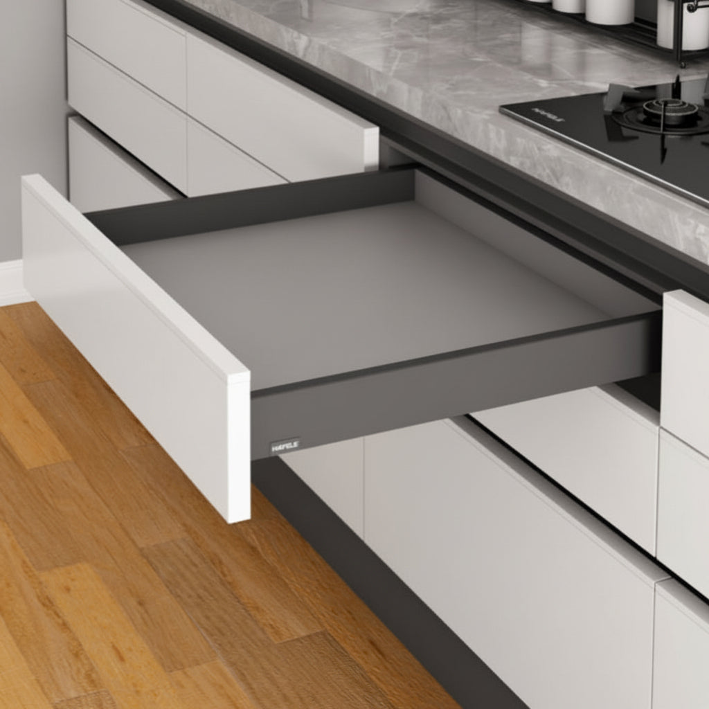 An open Hafele slim double-wall kitchen drawer featuring a matte anthracite grey finish and minimalist straight-line design. The drawer is integrated into white handle-less cabinetry under a grey marble countertop, showcasing high-end German engineering for modern kitchen organization.