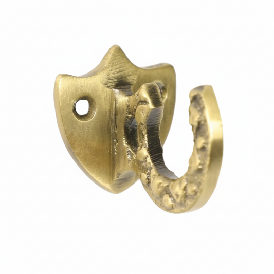 A decorative wall hook featuring a shield-shaped backplate and a textured curved hook in a brushed antique brass finish. The design includes two pre-drilled mounting holes for secure installation on walls, cabinets, or wardrobes.