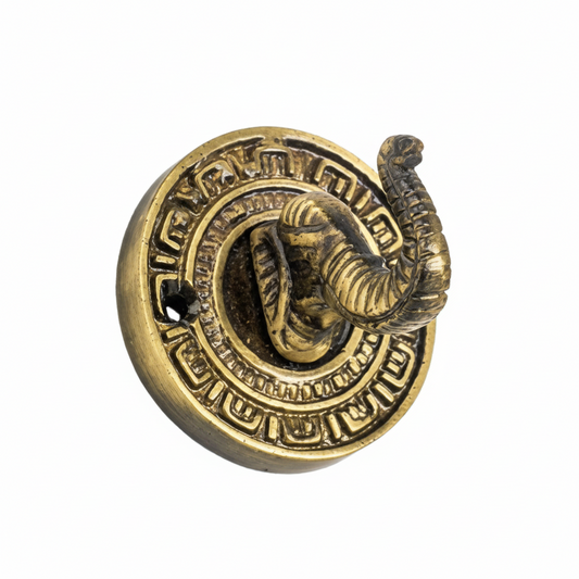 An ornate decorative wall hook featuring a detailed elephant head with an upturned trunk serving as the hook, set on a circular backplate with traditional geometric engravings in an antique brass finish.