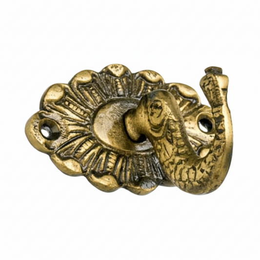 An ornate decorative wall hook featuring a detailed elephant head with an upturned trunk serving as the hook, mounted on a scalloped oval backplate with radial engravings and an antique brass finish. The piece includes two pre-drilled holes for secure wall mounting.