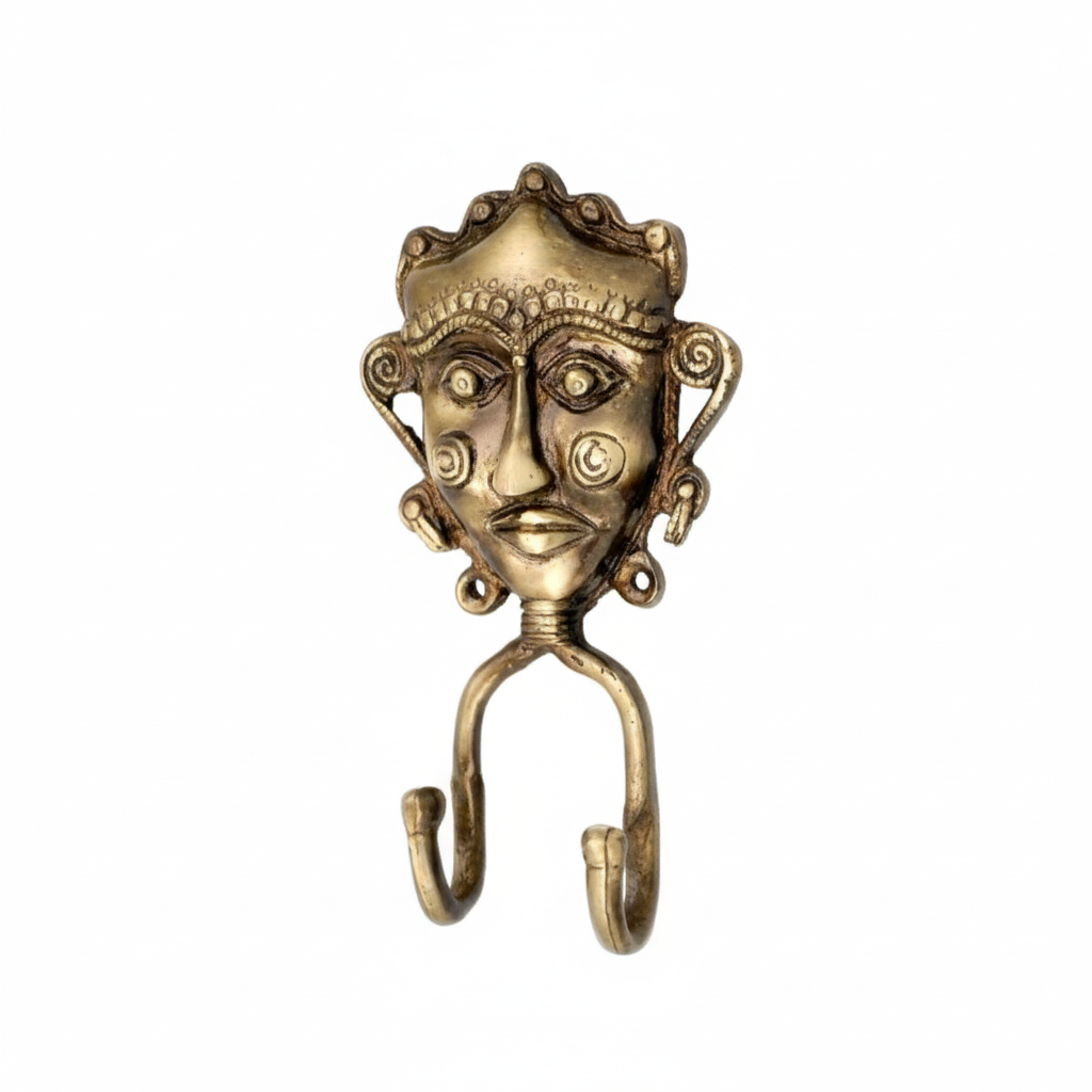 An antique brass double wall hook featuring a handcrafted Dhokra tribal mask with an ornate crown, spiral ear details, and engraved circular patterns on the cheeks. The decorative metal hook includes two curved hanging points, perfect for organizing keys or jewelry in a traditional Indian interior.