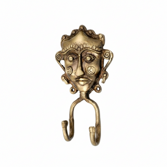 An antique brass double wall hook featuring a handcrafted Dhokra tribal mask with an ornate crown, spiral ear details, and engraved circular patterns on the cheeks. The decorative metal hook includes two curved hanging points, perfect for organizing keys or jewelry in a traditional Indian interior.
