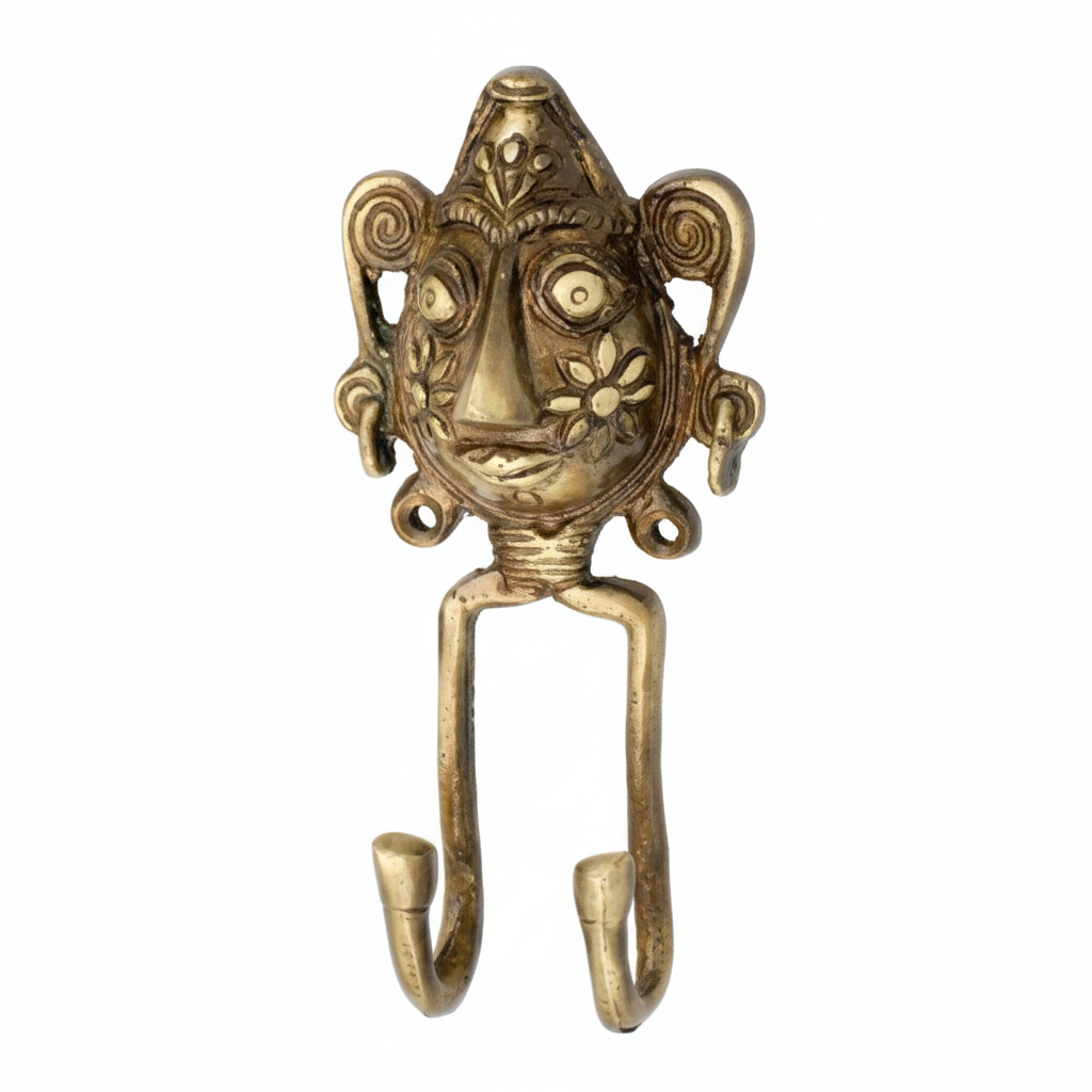 An antique brass double wall hook featuring a handcrafted Dhokra tribal face mask with intricate floral cheek engravings and spiral ear details. The decorative metal hook has two curved hanging points, ideal for organizing keys or jewelry in an ethnic-themed interior.