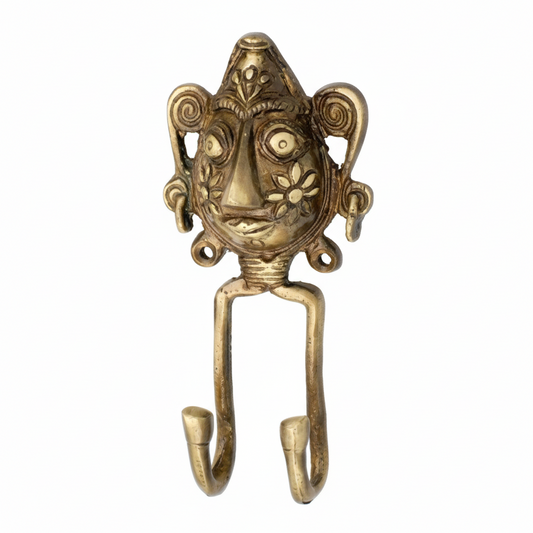An antique brass double wall hook featuring a handcrafted Dhokra tribal face mask with intricate floral cheek engravings and spiral ear details. The decorative metal hook has two curved hanging points, ideal for organizing keys or jewelry in an ethnic-themed interior.