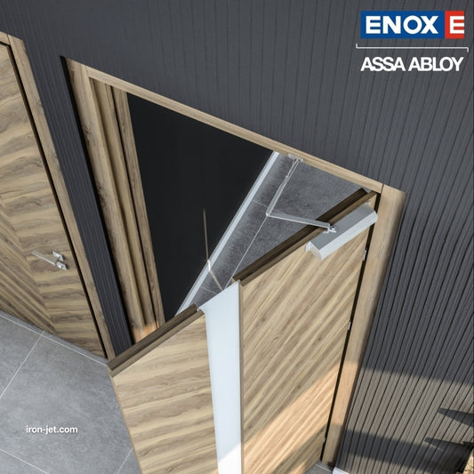 An installed ENOX (Assa Abloy) concealed overhead door closer on a modern light-wood grain interior door. The image showcases the hidden hydraulic mechanism and its sliding arm integrated into the door frame, maintaining a clean architectural aesthetic. This professional installation in a high-end office or residential setting demonstrates how concealed hardware provides smooth, controlled door closing without visible surface-mounted units.