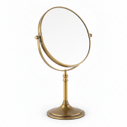 Grafiqe circular pedestal tabletop mirror in a premium antique brass finish, featuring a double-sided 360-degree swivel head for standard and magnified views on a slender stem and stable weighted base.