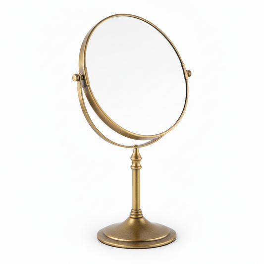 Grafiqe circular pedestal tabletop mirror in a premium antique brass finish, featuring a double-sided 360-degree swivel head for standard and magnified views on a slender stem and stable weighted base.