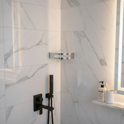 Lifestyle view of a Grafiqe stainless steel corner shower shelf installed on a white marble-tiled wall, positioned near a modern matte black shower fixture and a backlit vanity mirror for high-end bathroom organization.