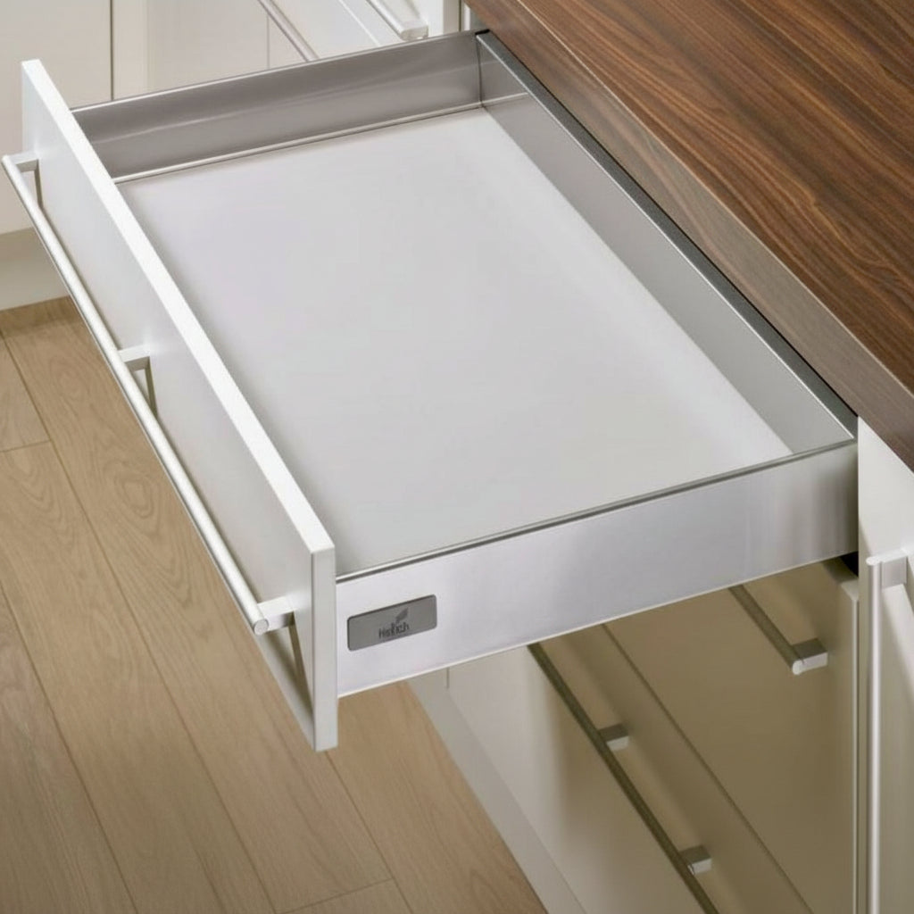 An open Hettich slim double-wall kitchen drawer featuring a premium silver metallic finish and a minimalist straight-line design. The drawer is integrated into white cabinetry under a natural wood countertop, showcasing high-end German engineering for modern kitchen interior organization.