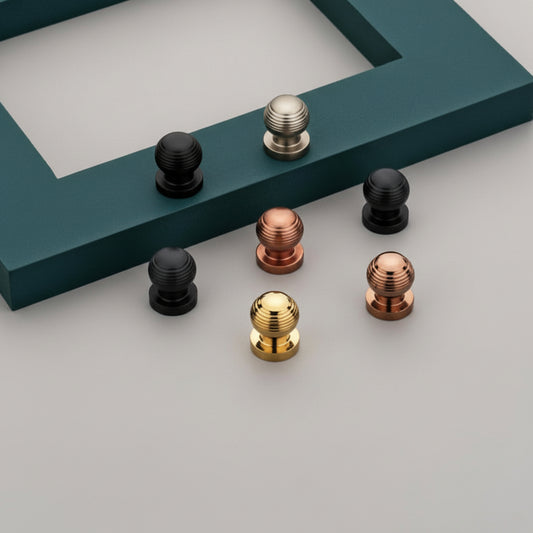 A variety of Creatick KB-02 modern ball-shaped cabinet knobs with a ribbed texture, available in matte black, satin gold, rose gold, and brushed gray finishes. These elegant furniture knobs are displayed on a gray surface with a teal decorative frame, offering a high-end hardware solution for kitchen cabinets, drawers, and wardrobe doors.