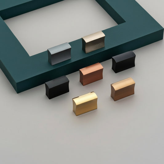 A collection of Creatick KB-36 modern rectangular cabinet knobs with a sleek, concave profile. These contemporary furniture pulls are displayed in various finishes, including matte black, polished gold, rose gold, and brushed silver, arranged on a grey surface with a teal accent frame—ideal for upscale kitchen cabinets and minimalist drawer hardware.
