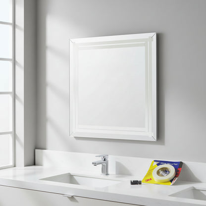 A modern bathroom scene showing a large square mirror mounted on a gray wall above a white vanity and sink. The 3M Mirror Mount Tape packaging and primer are visible on the counter, demonstrating a successful drill-free mirror installation.